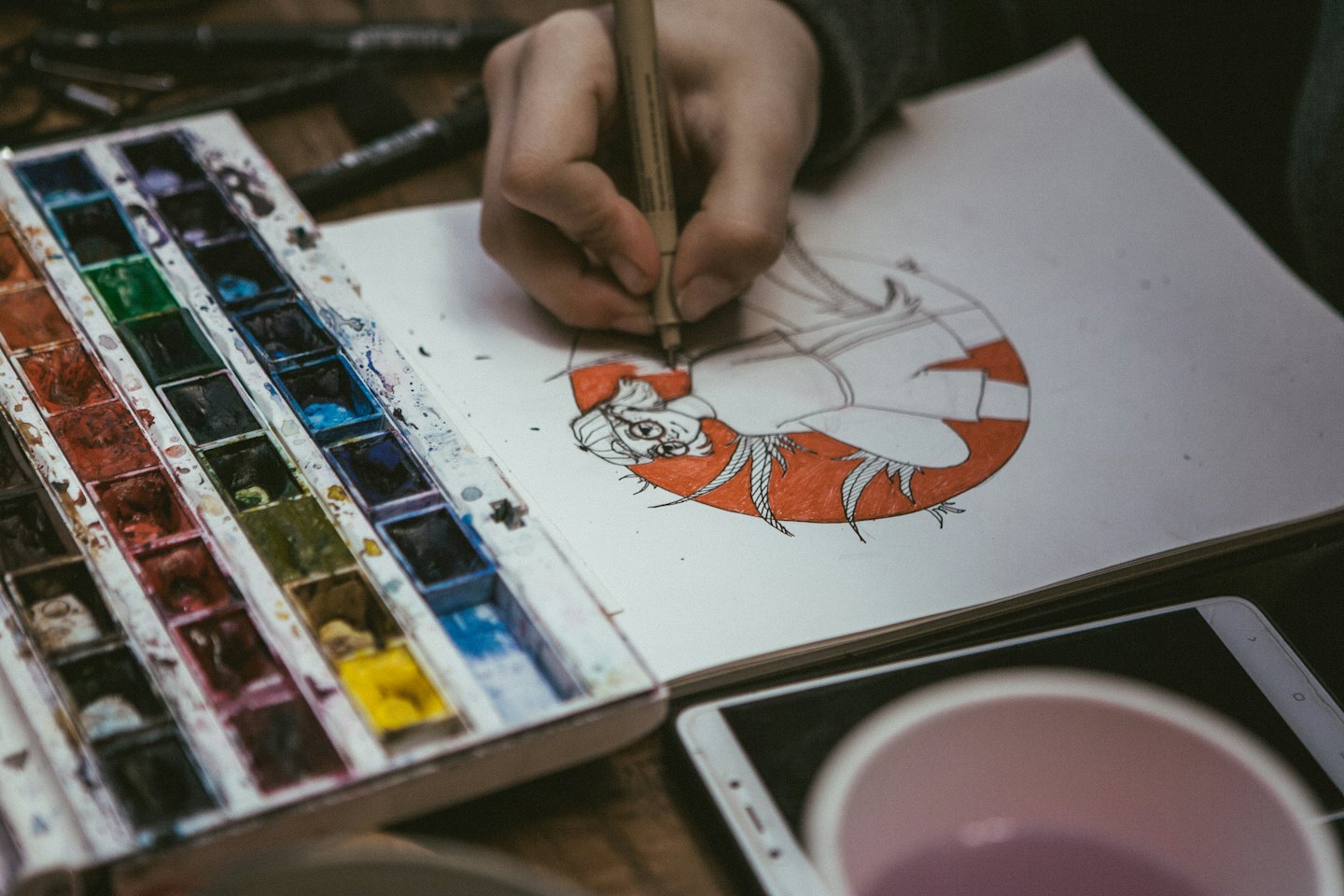 Artista che disegna un personaggio anime su un taccuino vicino a delle tempere.