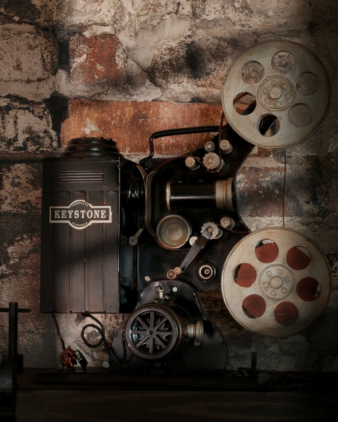 Proiettore cinematografico vintage Keystone con bobine di pellicola su un muro di mattoni.